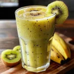 Smoothie de kiwi e banana em copo transparente com pedaços de fruta ao redor.