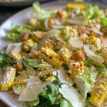 ['Salada Caesar de verão com milho grelhado em close.', 'Porção de salada Caesar com alface, milho e parmesão.', 'Salada Caesar fresca com milho charred e molho cremoso.', 'Vista lateral de salada Caesar de verão com ingredientes coloridos.']