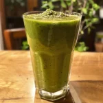 ['Smoothie verde em um copo, com couve, maçã e gengibre.', 'Close de um smoothie saudável verde com ingredientes frescos.', 'Bebida verde em copo, mostrando textura cremosa e ingredientes.', 'Smoothie de couve, maçã e gengibre em uma mesa.']