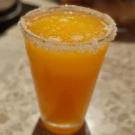 ['Copo com suco de tangerina fresco e gelo.', 'Suco de tangerina servido em copo com gelo.', 'Close de um copo de suco de tangerina refrescante.', 'Bebida de suco de tangerina com cubos de gelo.']