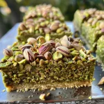 ['Bolo vegano de pistache com cobertura cremosa.', 'Fatia de bolo vegano de pistache em close.', 'Vista lateral do bolo vegano de pistache.', 'Bolo de pistache vegano decorado.']