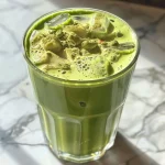 ['Iced Matcha Latte em um copo com gelo e espuma de baunilha.', 'Close-up de um Iced Matcha Latte com purê de morango.', 'Iced Matcha Latte servido com cubos de gelo e detalhes de espuma.', 'Bebida verde refrescante com matcha e creme espumoso.']