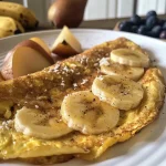 ['Omelete de banana e aveia com frutas frescas ao lado.', 'Close de uma omelete de banana e aveia servida em um prato.', 'Detalhe de uma omelete de aveia com banana e cobertura de frutas.', 'Omelete de banana e aveia com coco e mel decorativa.']
